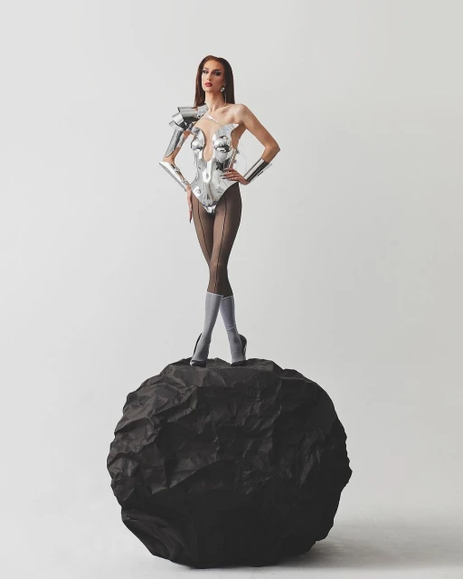 A slender woman with long brown hair, wearing a futuristic silver bodysuit and heels, poses confidently on a black rock in a studio setting.