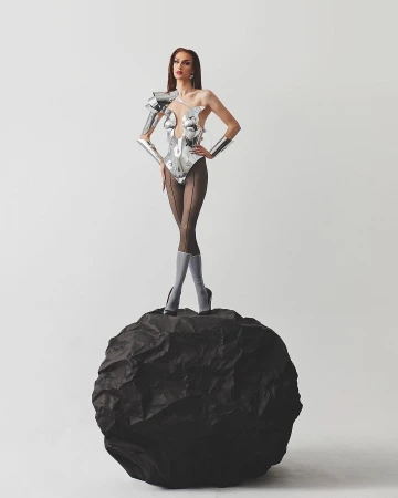 A slender woman with long brown hair, wearing a futuristic silver bodysuit and heels, poses confidently on a black rock in a studio setting.