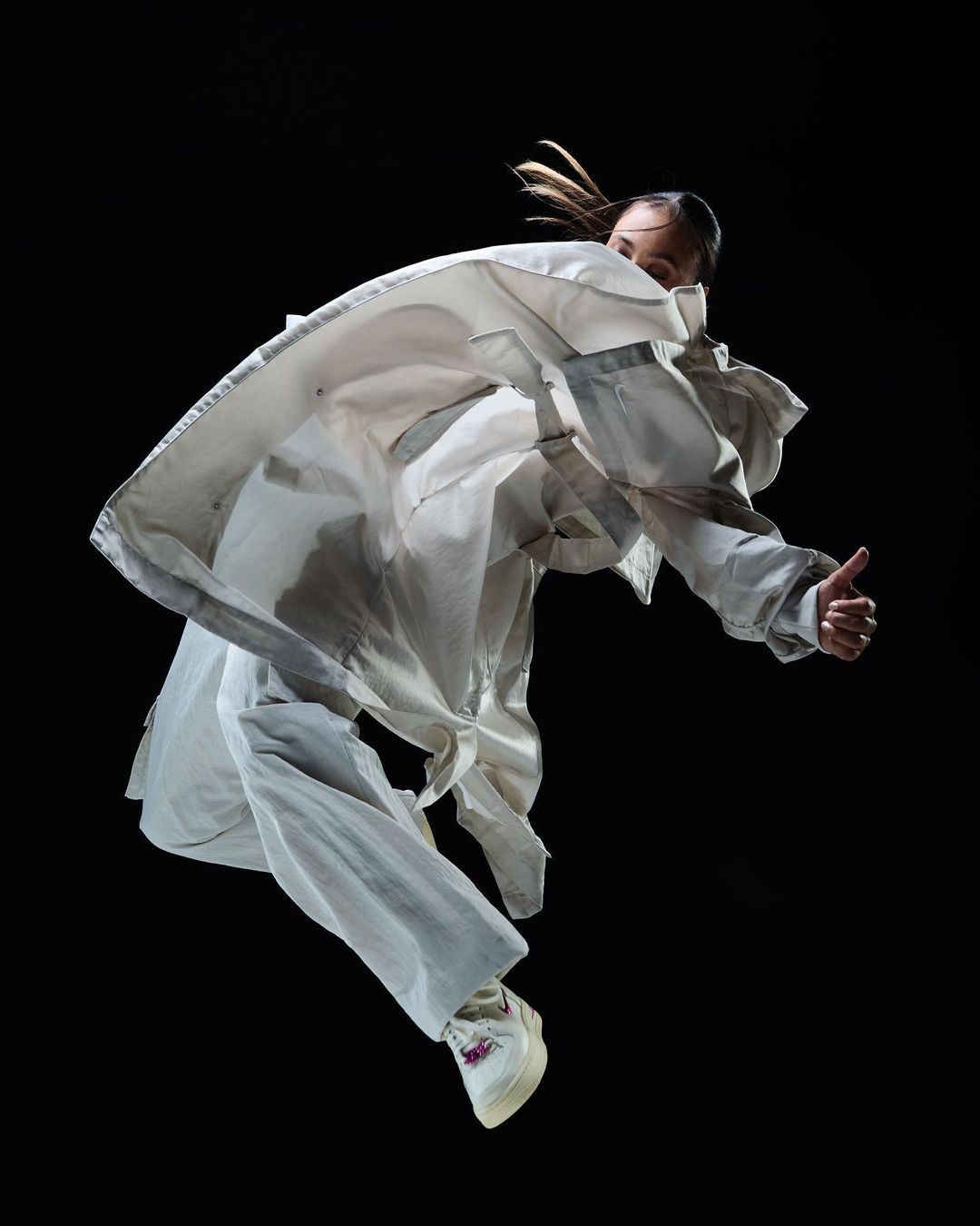 Woman in white oversized jacket and pants leaps in mid-air with a thumbs-up against a dark backdrop, motion and energy.