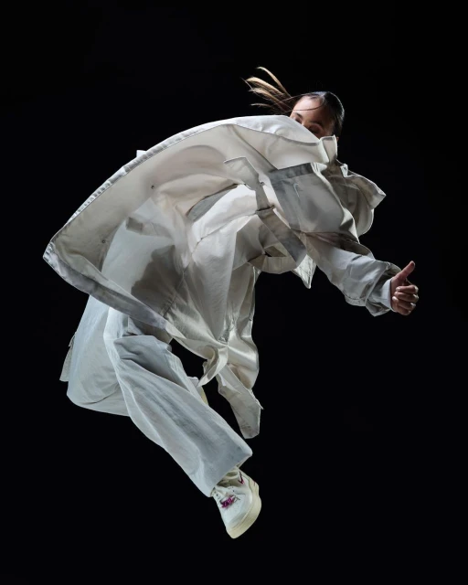 Woman in white oversized jacket and pants leaps in mid-air with a thumbs-up against a dark backdrop, motion and energy.
