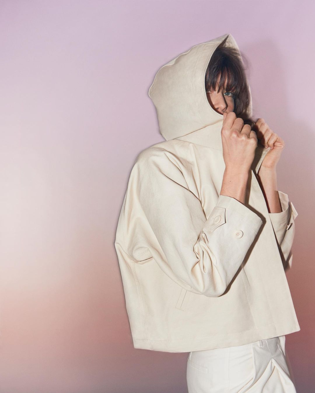 Woman in a hooded jacket, looking over her shoulder against a gradient backdrop of peach and lavender, fashion photography.