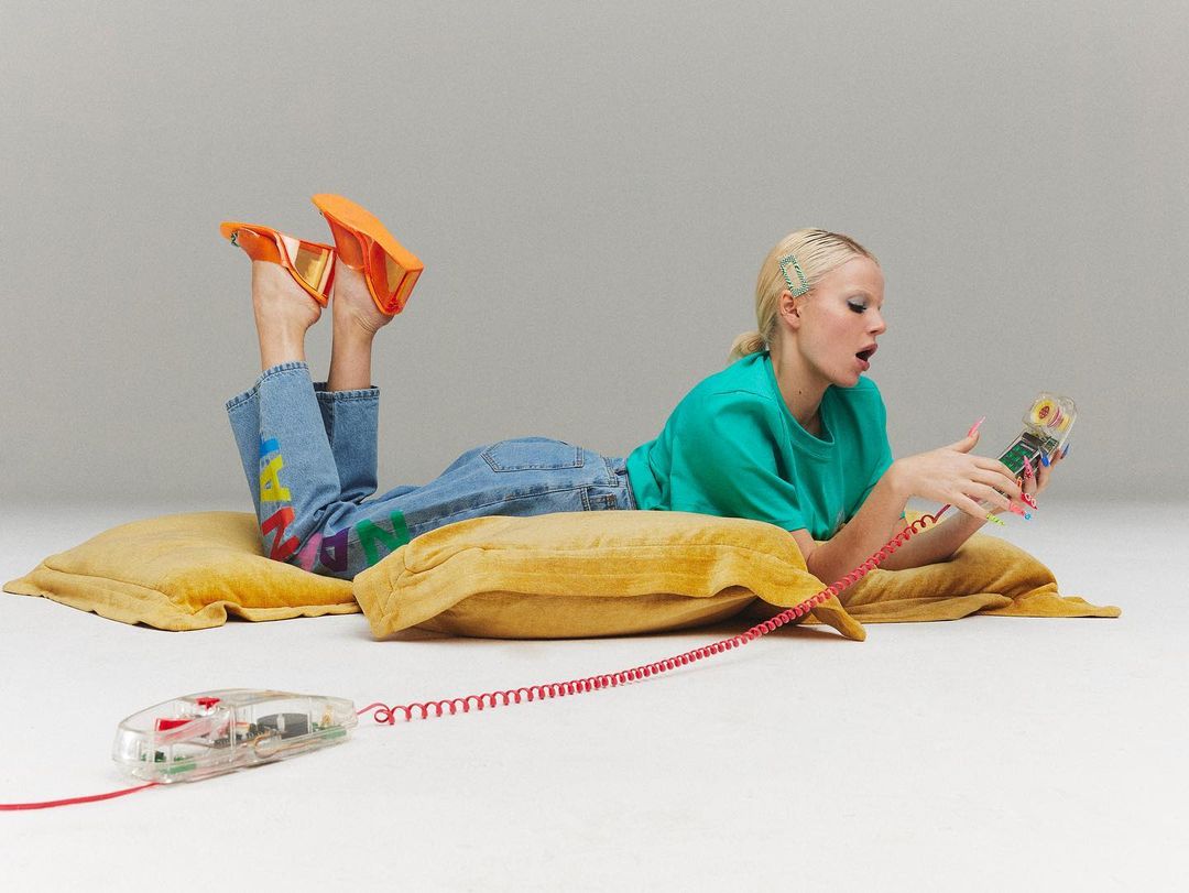Woman lying on pillows using a retro clear telephone, wearing colorful jeans and orange heels against a neutral background.
