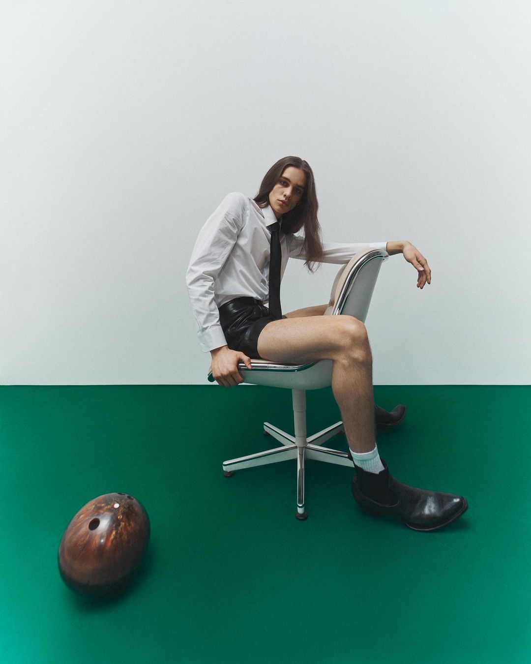 Man in a white shirt and black tie seated on an office chair, with a bowling ball on the green and white floor in a studio.