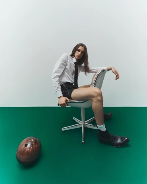 Man in a white shirt and black tie seated on an office chair, with a bowling ball on the green and white floor in a studio.