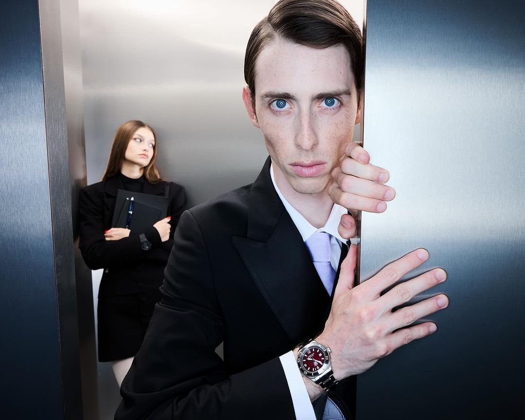 Indoor shot of a businessman looking out from behind a metal panel, with a businesswoman in the background holding a file.