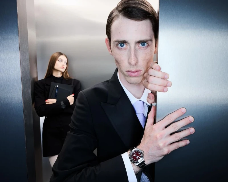 Indoor shot of a businessman looking out from behind a metal panel, with a businesswoman in the background holding a file.