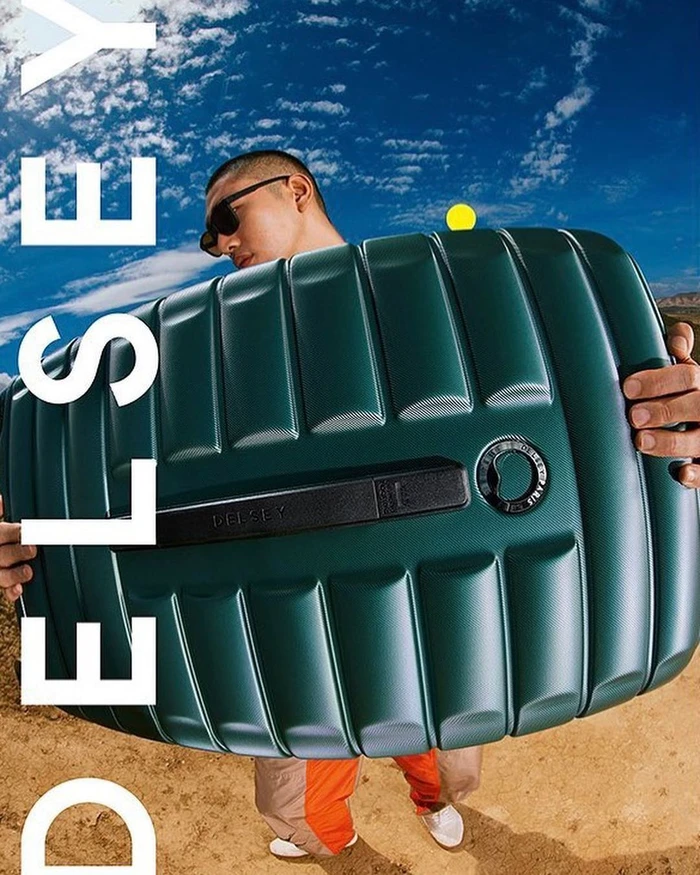 Man holding a large green ribbed hardshell suitcase against a blue sky with clouds and sandy ground.