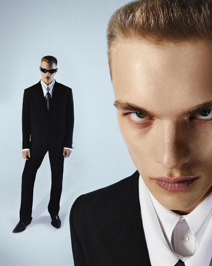 Extreme close-up portrait of a man in a black suit with a second full-length figure standing in the background against a light backdrop