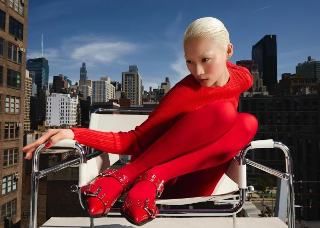Model with bleached blonde hair wearing bright red ribbed top and stockings seated in a modern white armchair outdoors on a rooftop