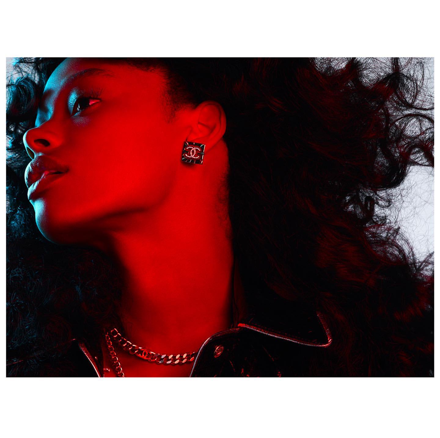 Profile shot of a woman with curly dark hair wearing a black leather jacket and gold chain. Red and blue lighting illuminates the scene.