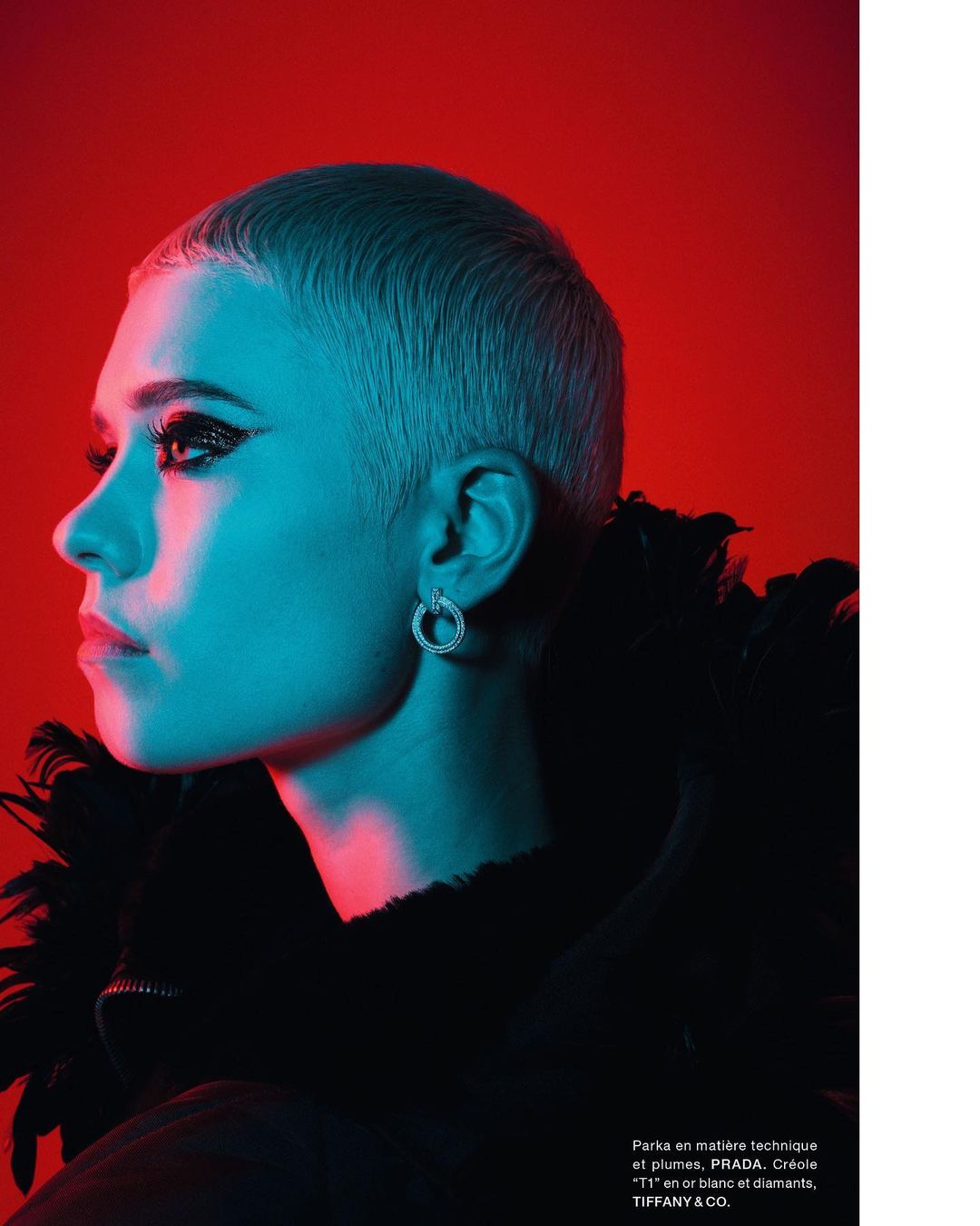 Profile of a woman with short blonde hair, wearing earrings and a black feathered jacket against a red backdrop with blue lighting.