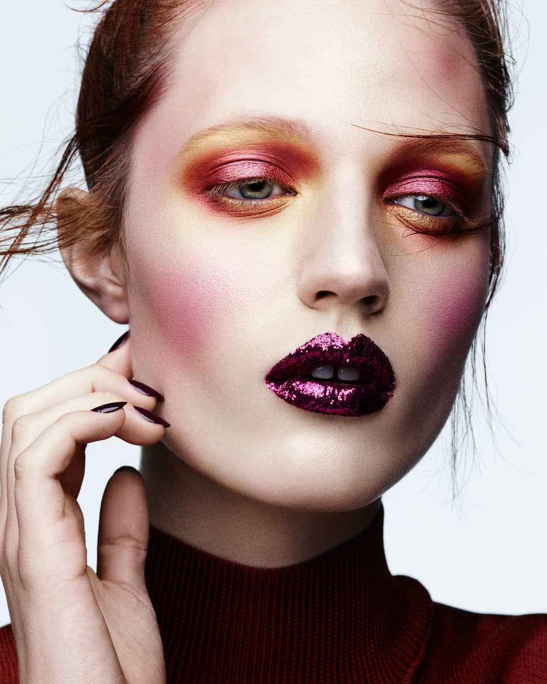 Close-up portrait of a fair-skinned woman with red hair and dramatic makeup, featuring glittery lips and colorful eyeshadow against a light background.