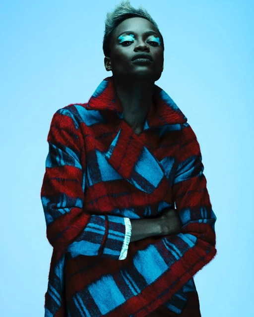 Fashion portrait of an African American woman in a blue and red plaid coat with blue eyeshadow, posing against a light blue background.