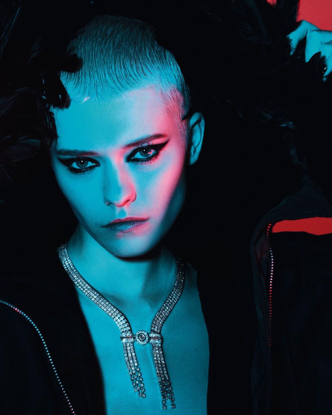 Close-up portrait of a model with a buzzcut wearing a diamond necklace and dark eyeliner under blue and red lighting.