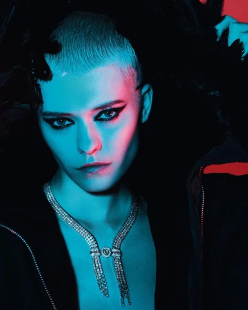 Close-up portrait of a model with a buzzcut wearing a diamond necklace and dark eyeliner under blue and red lighting.