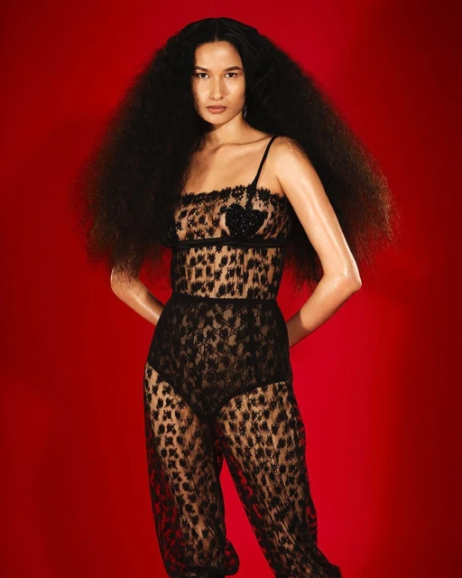 A woman with big, curly dark hair poses against a red background, wearing a black lace animal-print top and matching pants.