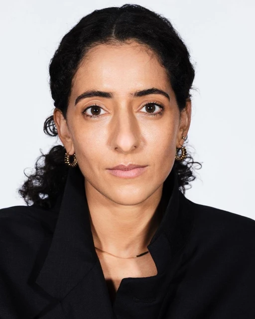 Portrait of a woman with dark curly hair and a serious expression, wearing a black jacket, against a white background in a studio setting.