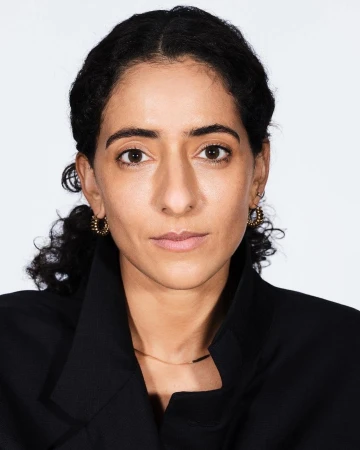 Portrait of a woman with dark curly hair and a serious expression, wearing a black jacket, against a white background in a studio setting.