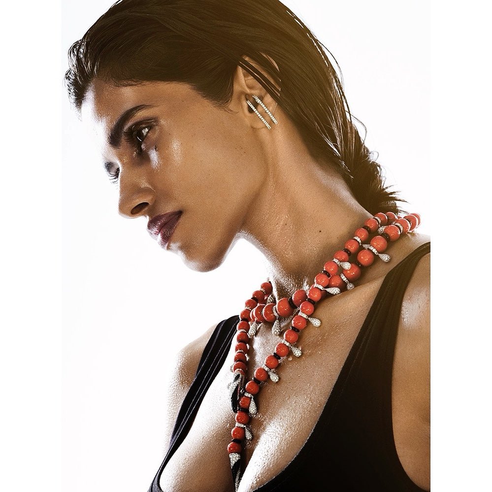 Profile of a woman with damp dark hair wearing red and white beaded necklace with a black top against a white background.