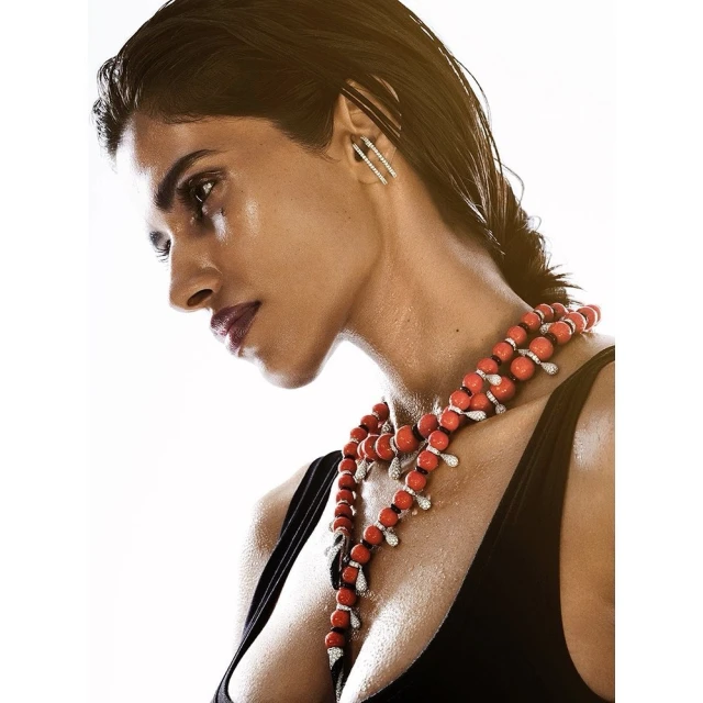 Profile of a woman with damp dark hair wearing red and white beaded necklace with a black top against a white background.