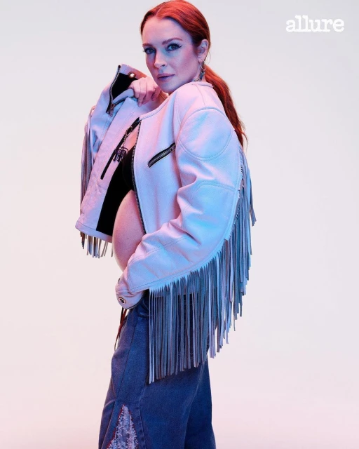 Red-haired woman poses in jeans and a white fringe jacket for an Allure magazine shot in a studio with diffused lighting.