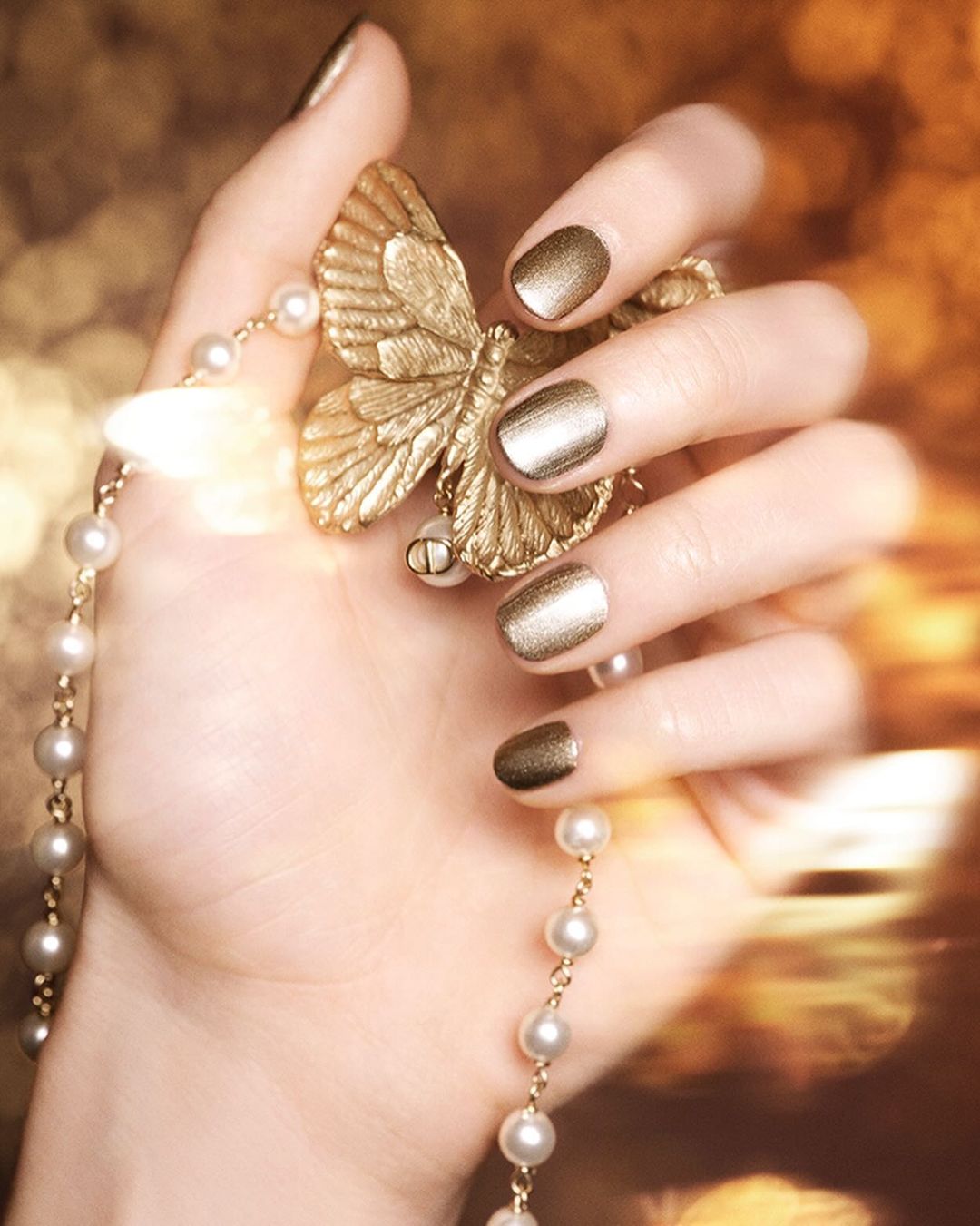 Close-up of a hand with gold metallic nails holding a gold butterfly and a pearl necklace in a soft-focus, warm-toned setting.