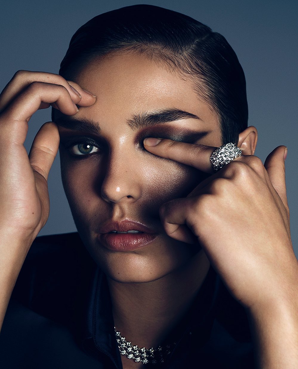 Portrait of a brunette adult female with hand near face wearing diamond ring and necklace against a grey backdrop in a studio setting.