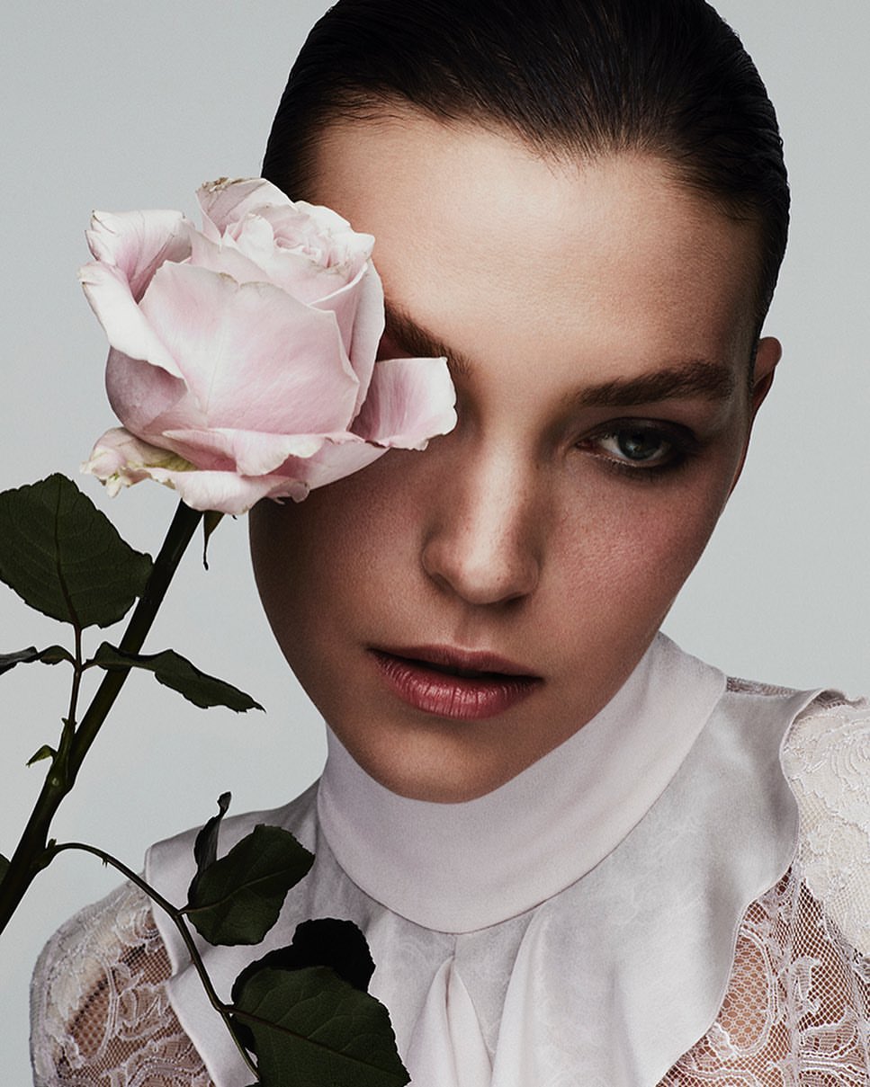 Portrait of a fair-skinned woman with a light pink rose covering one eye, wearing a white high-collared lace shirt. Studio lighting on a grey background.