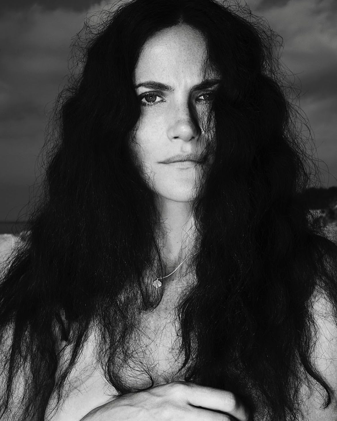 Striking black and white portrait of a woman with long, dark, curly hair against a cloudy sky, creating a dramatic, natural aesthetic.