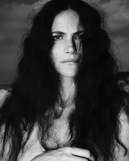 Striking black and white portrait of a woman with long, dark, curly hair against a cloudy sky, creating a dramatic, natural aesthetic.