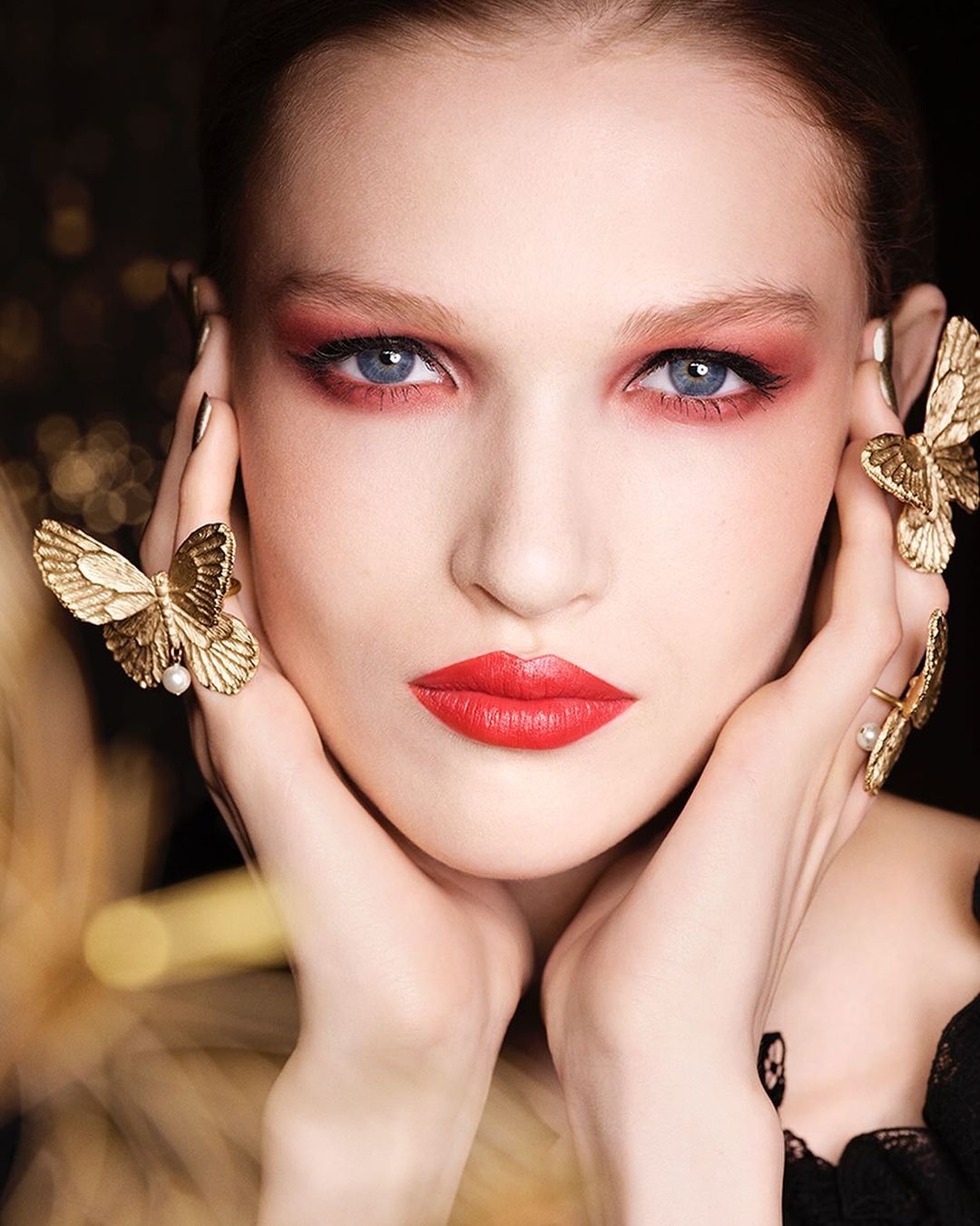 Close-up of a woman with red eye shadow and lipstick wearing butterfly jewelry posing in front of a dark background.