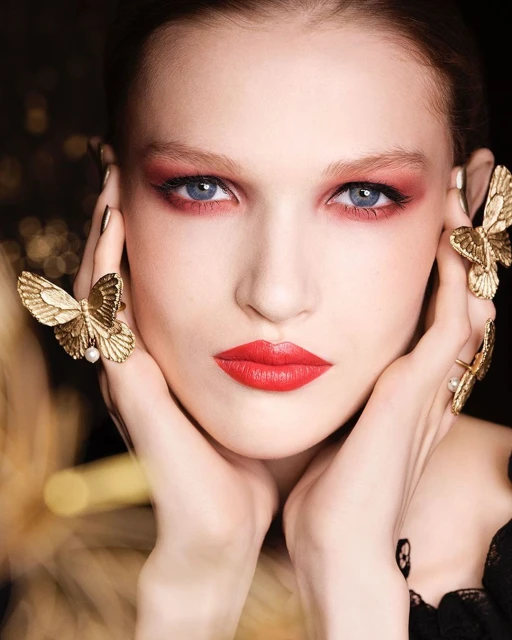 Close-up of a woman with red eye shadow and lipstick wearing butterfly jewelry posing in front of a dark background.