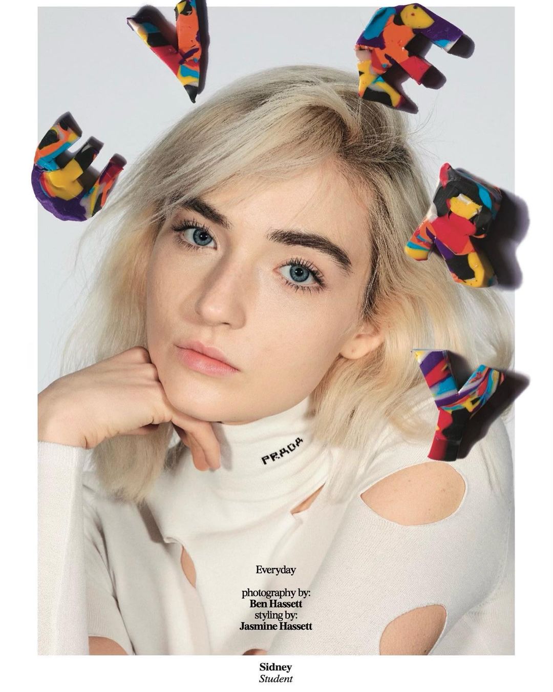 Close-up of a blonde woman wearing a white long-sleeve shirt with cut-outs, posing with colorful clay shapes surrounding her head.