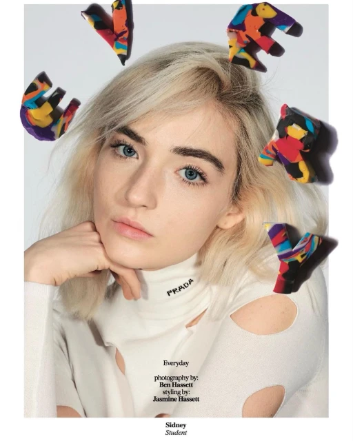 Close-up of a blonde woman wearing a white long-sleeve shirt with cut-outs, posing with colorful clay shapes surrounding her head.