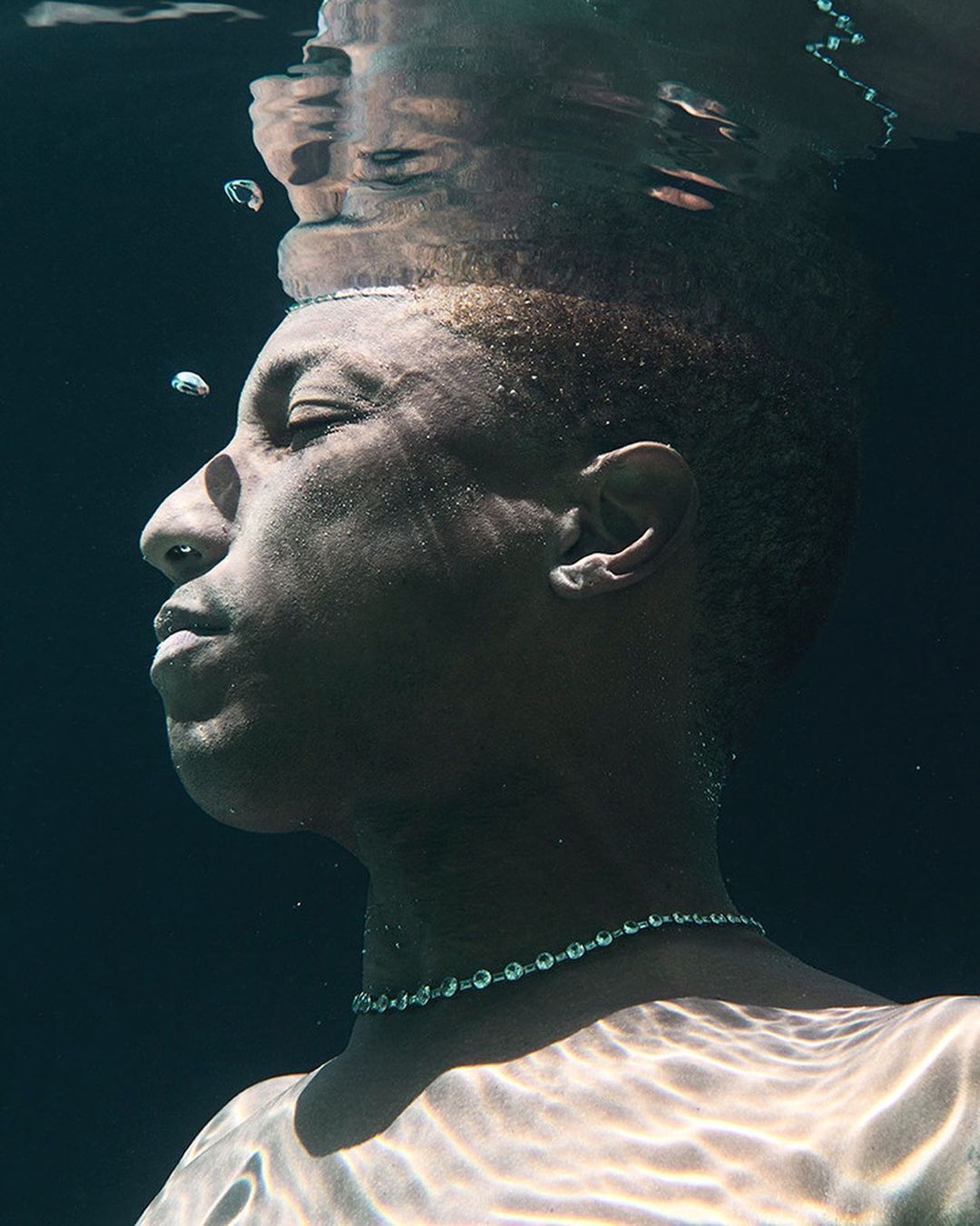 Underwater portrait of an African American male with short blond hair. He is wearing a beaded necklace, and his head is partially submerged.