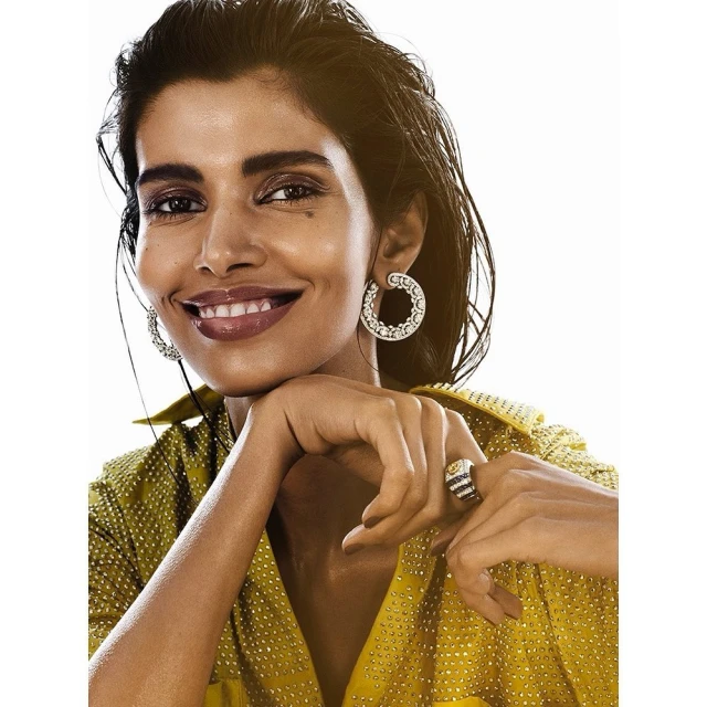 Close-up studio shot of a smiling adult woman with dark hair and diamond jewelry, wearing a yellow, studded top against a white background.