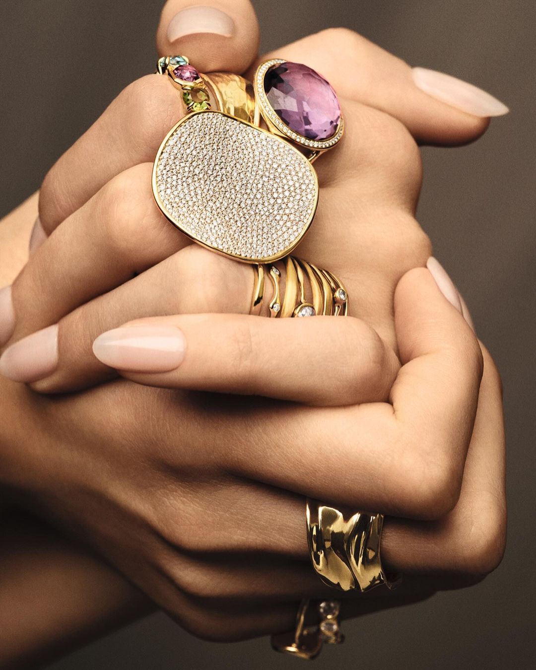 Close-up of a person's hands adorned with multiple statement rings in gold with gemstones, set against a neutral-toned background.
