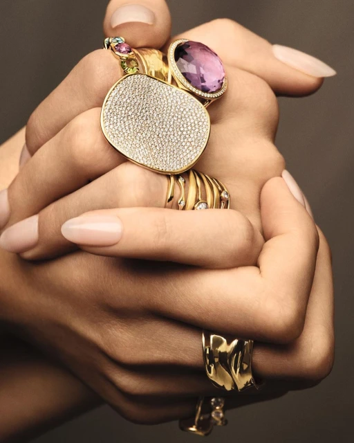 Close-up of a person's hands adorned with multiple statement rings in gold with gemstones, set against a neutral-toned background.