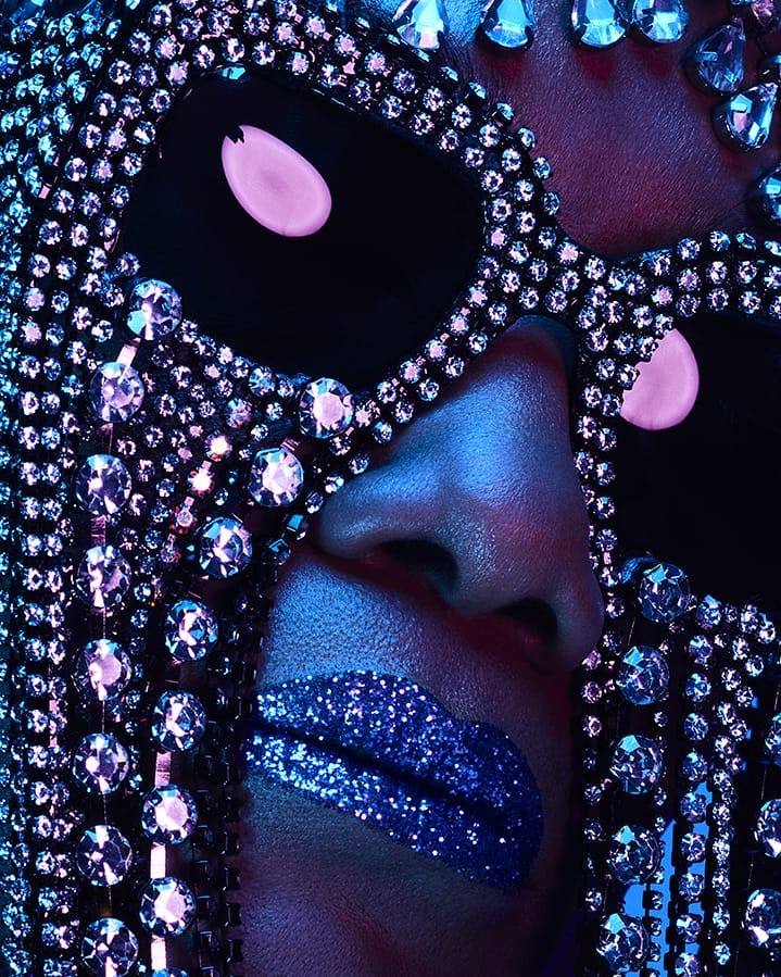 Close-up portrait of a woman with bright blue glitter lips and a jeweled mask covering the top half of her face, dramatic lighting.