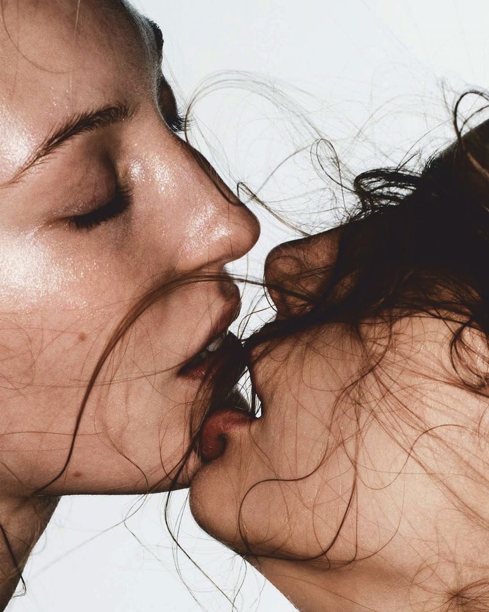 Extreme close-up portrait of two women kissing intimately with loose strands of hair framing their faces against a plain background