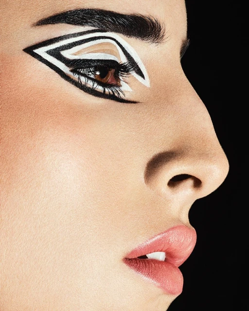 Close-up studio shot of a woman's face with graphic black and white eye makeup, soft pink lips, dark hair, and a black background.