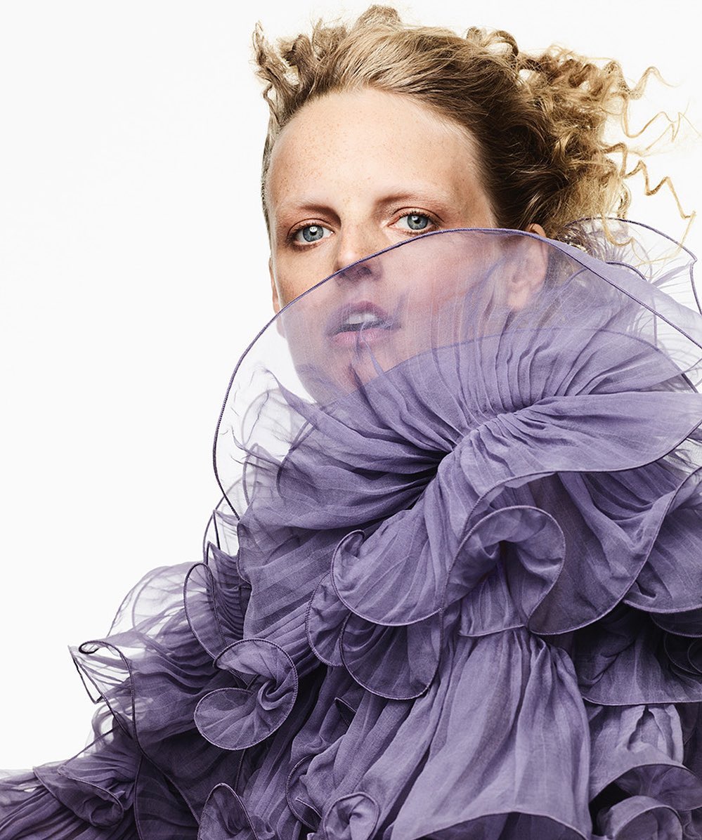 Fashion portrait of a blonde woman with blue eyes wearing a lilac ruffled top against a white background in a studio setting.