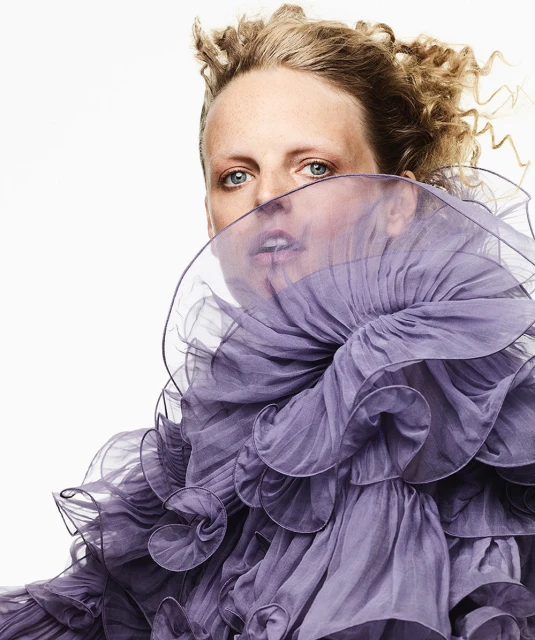 Fashion portrait of a blonde woman with blue eyes wearing a lilac ruffled top against a white background in a studio setting.