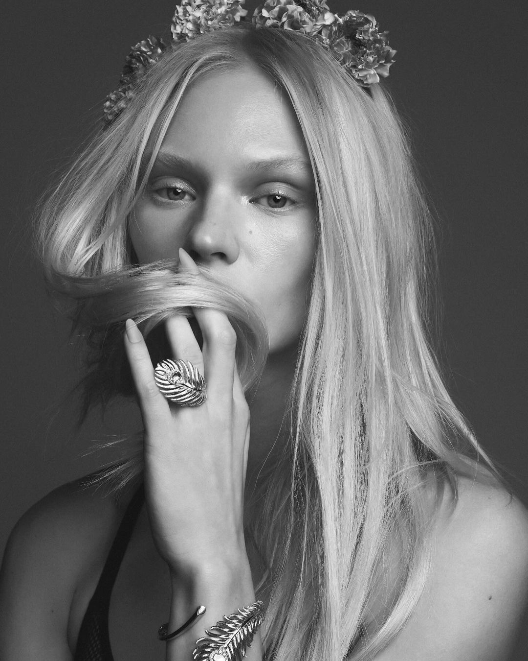 Black and white portrait of a woman with long blond hair, flower crown, and jewelry posing in front of a neutral background.
