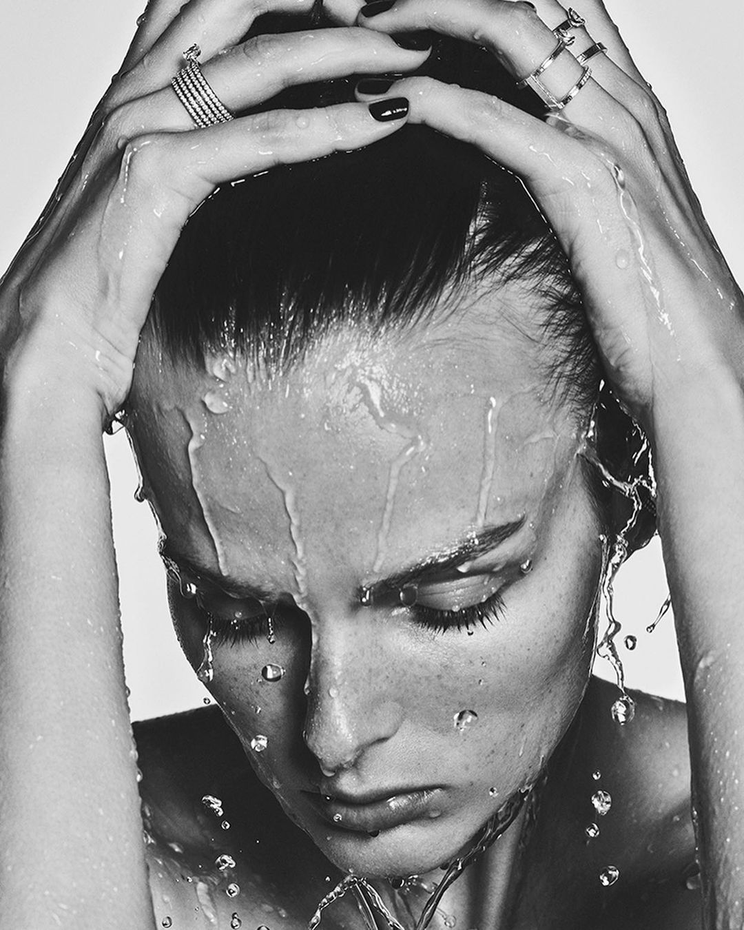 Close-up black and white portrait of a woman with water cascading over her face and hands covering her hair, showcasing a contemplative mood.