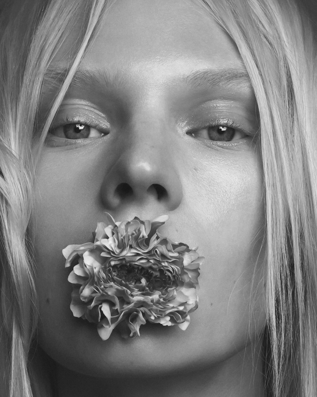 Close-up studio shot of a fair-skinned adult woman with blonde hair, holding a flower in her mouth, in a high-key black-and-white image.