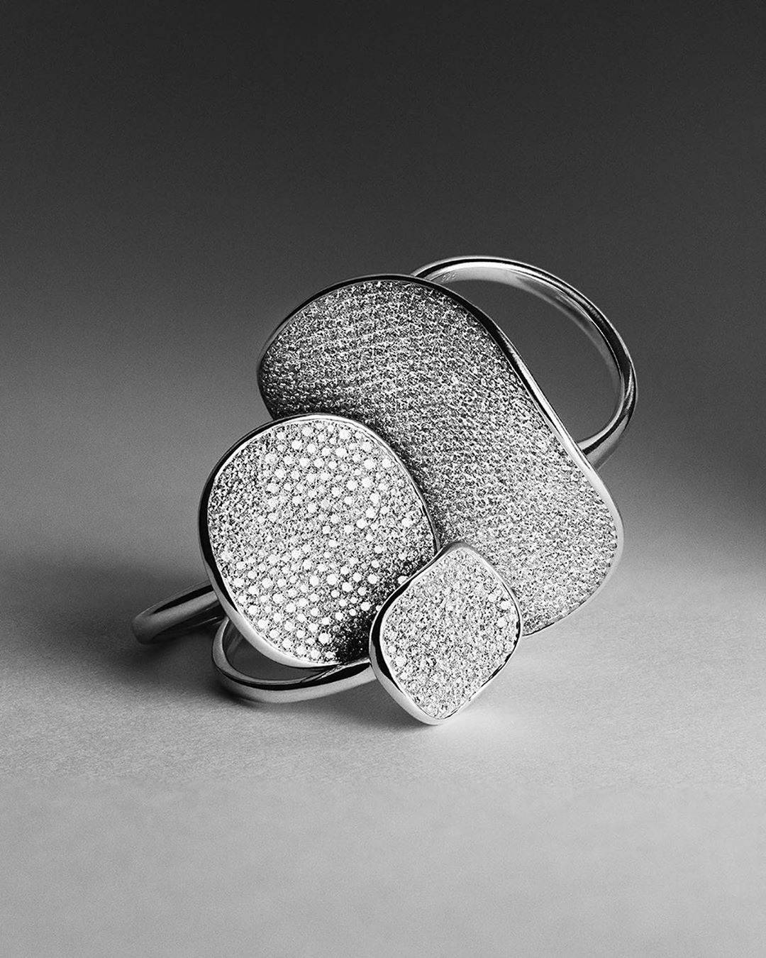 Close-up of a textured, geometric silver ring featuring a square, oval, and organic shape, set against a neutral gray gradient background.