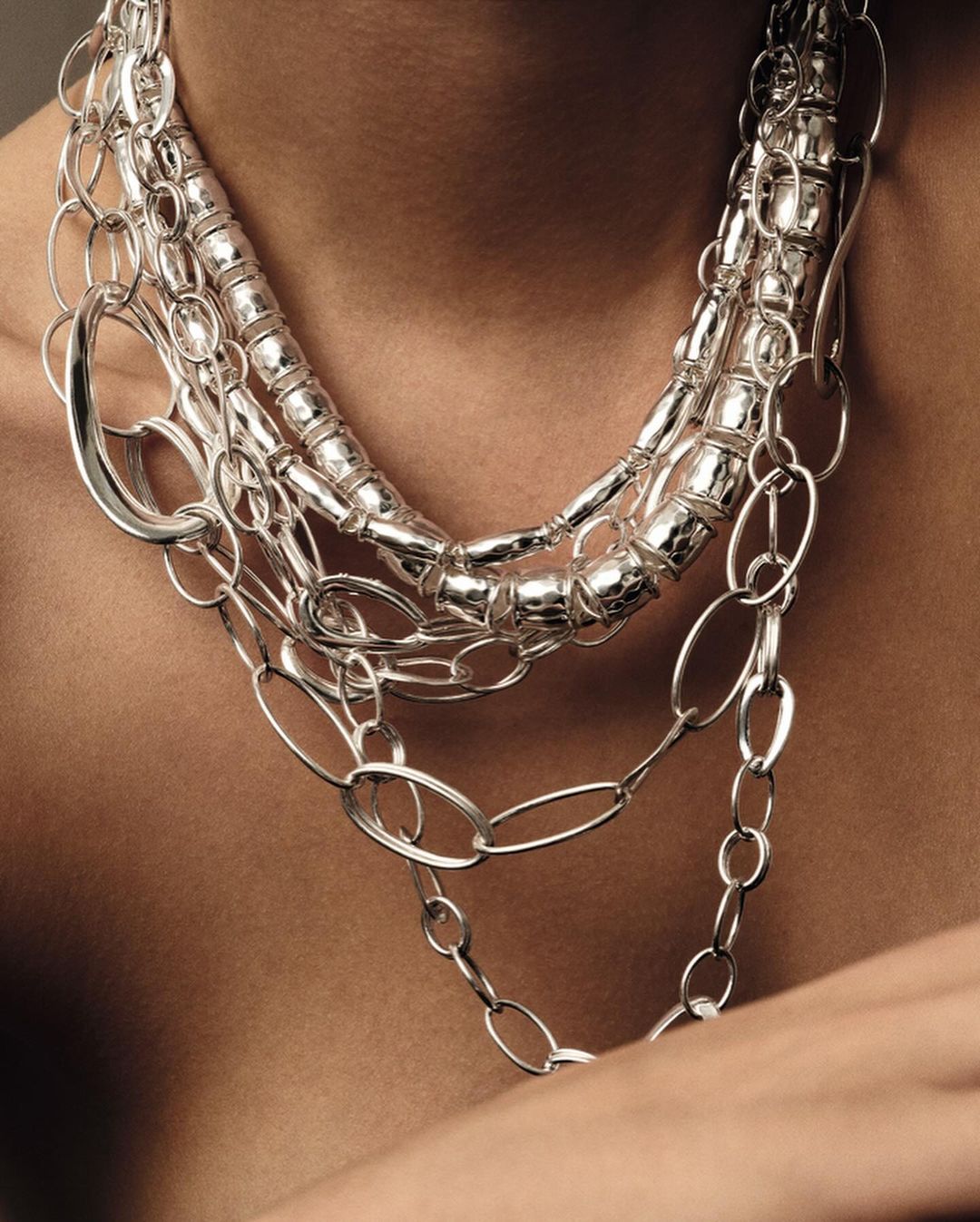 Close-up shot of a person wearing a multi-strand silver necklace, featuring various chain styles and metallic textures, against a neutral backdrop.