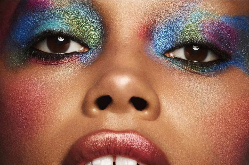 Close-up of a woman's face with colorful makeup. Eyes are adorned with blue, green, and purple eyeshadow. Pink blush highlights cheeks.
