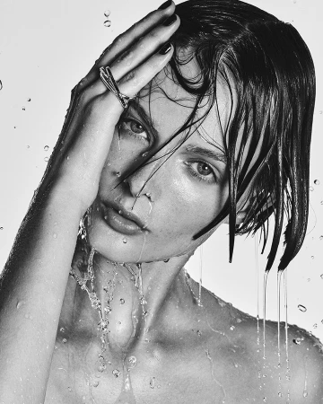 Close-up of a woman with wet hair and skin, wearing a metallic ring, photographed against a white background by Ben Hassett.